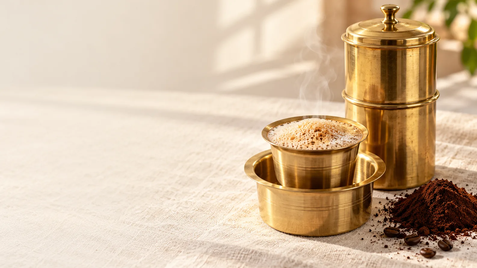 A brass filter, tumbler and dabarah of frothy coffee on linen beside a heap of ground coffee