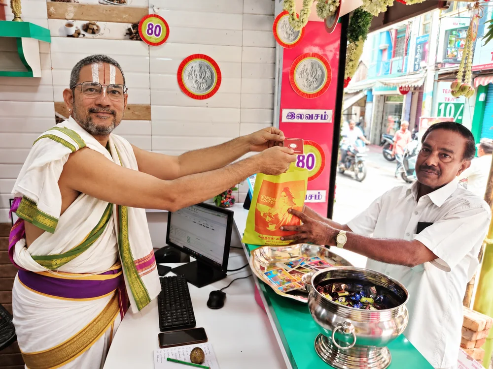 Rajagopalan N. behind his counter, handing a packet to a customer, the street visible beyond